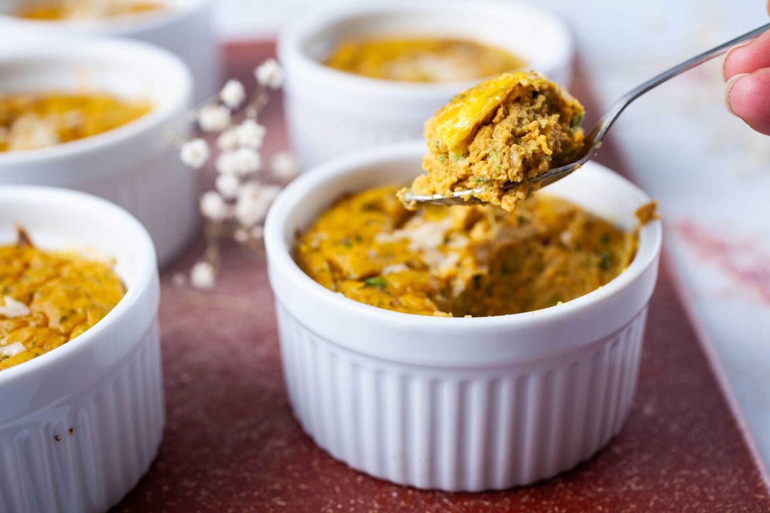A close-up of individual white ramekins filled with a baked, golden brown savory dish, perfect for pumpkin season, with a spoon lifting a portion from one ramekin.