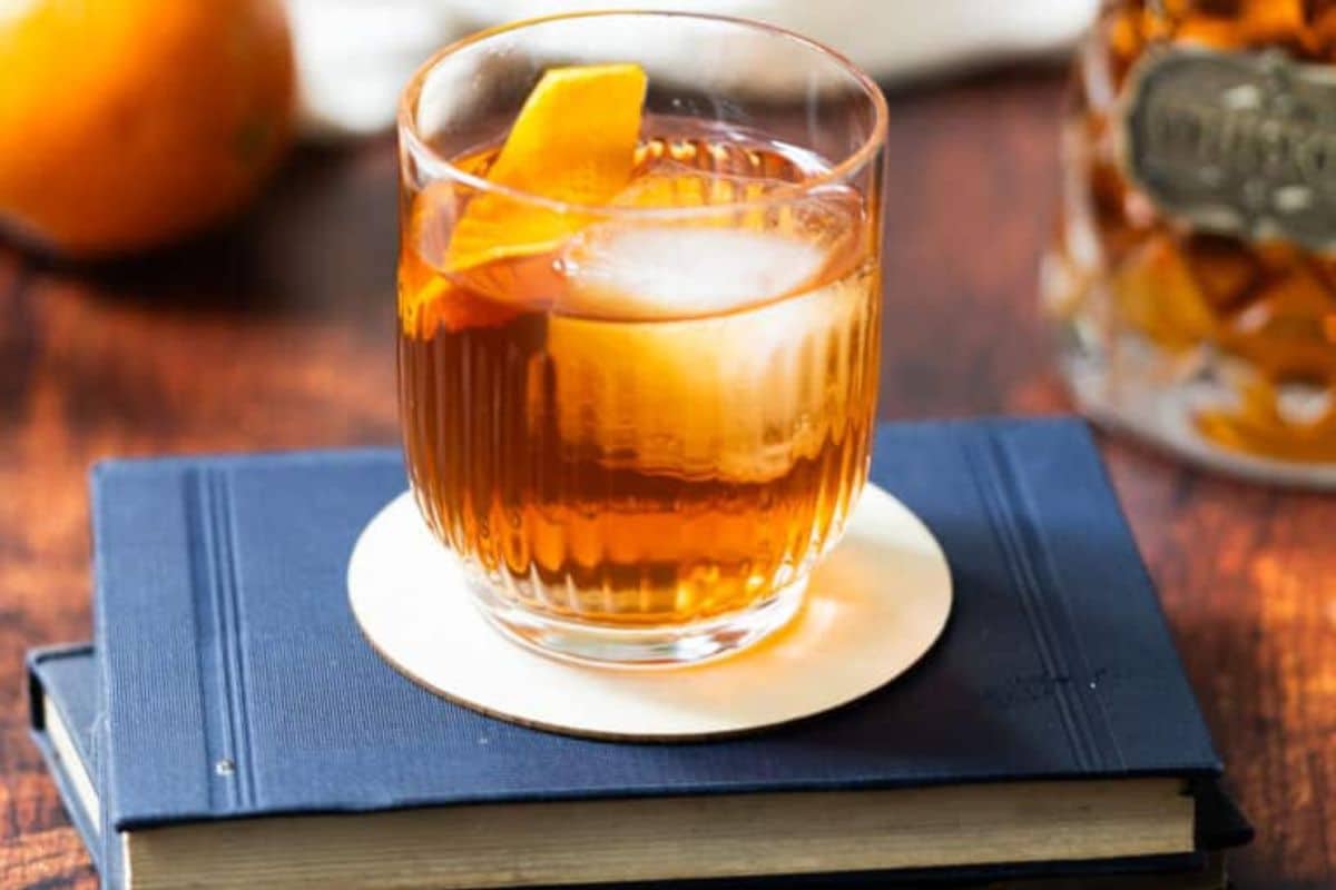 A glass of amber-colored cocktail with ice and an orange peel sits on a coaster atop a closed blue book, perfect for sipping by the fire, with a jar and an orange blurred in the background.