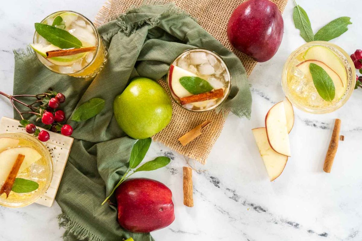 Overhead view of apple-themed drinks with ice, garnished with cinnamon sticks and sage leaves, surrounded by apples, apple slices, berries, and green fabric on a marble surface—perfect inspiration for celebrating apple season with delicious seasonal recipes.