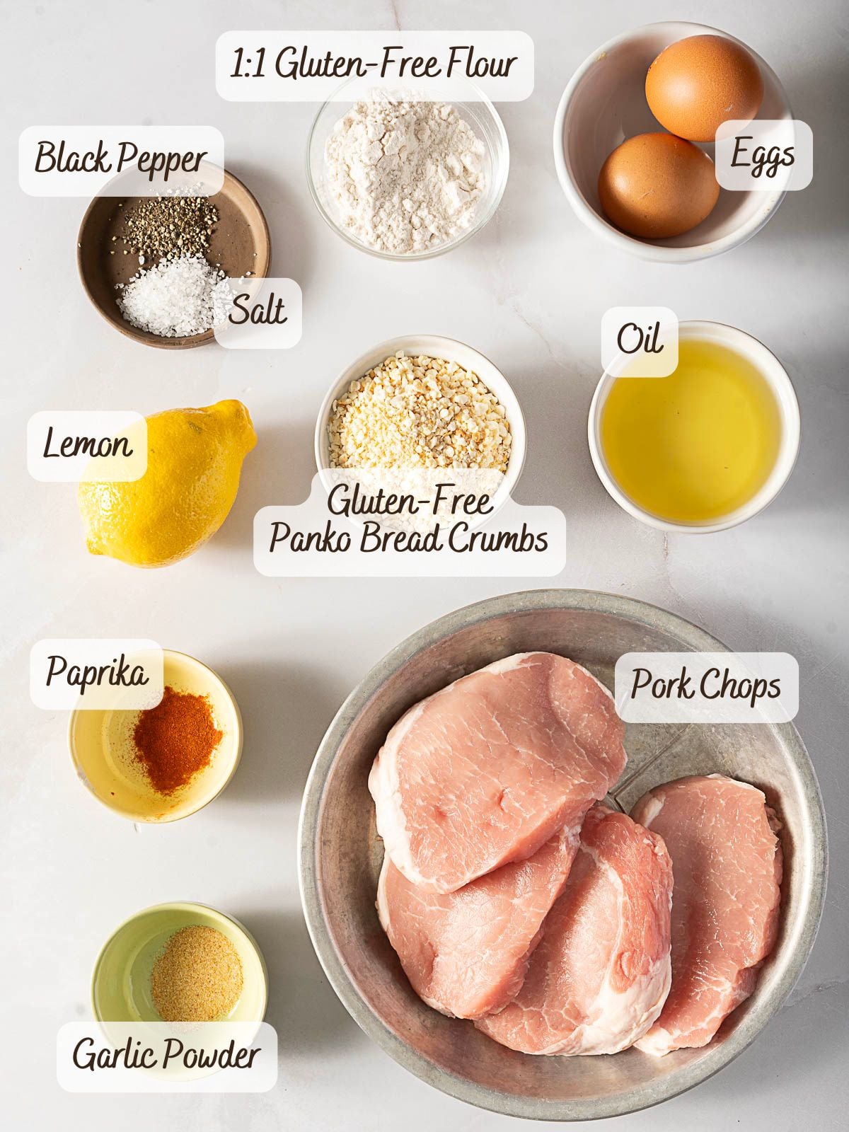 Raw pork chops, eggs, oil, gluten-free bread crumbs, gluten-free flour, lemon, salt, black pepper, paprika, and garlic powder are arranged separately on a white surface.