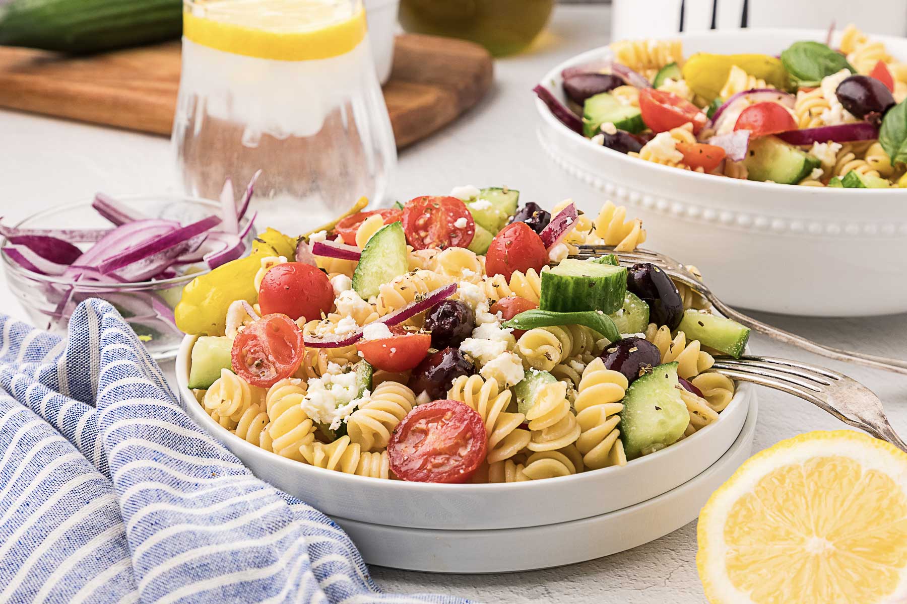 A plate of rotini pasta salad with cherry tomatoes, cucumbers, red onions, olives, feta cheese, and peppers sits next to a glass of water with lemon—one of those perfect potluck salads that travel well and are always a hit at gatherings.