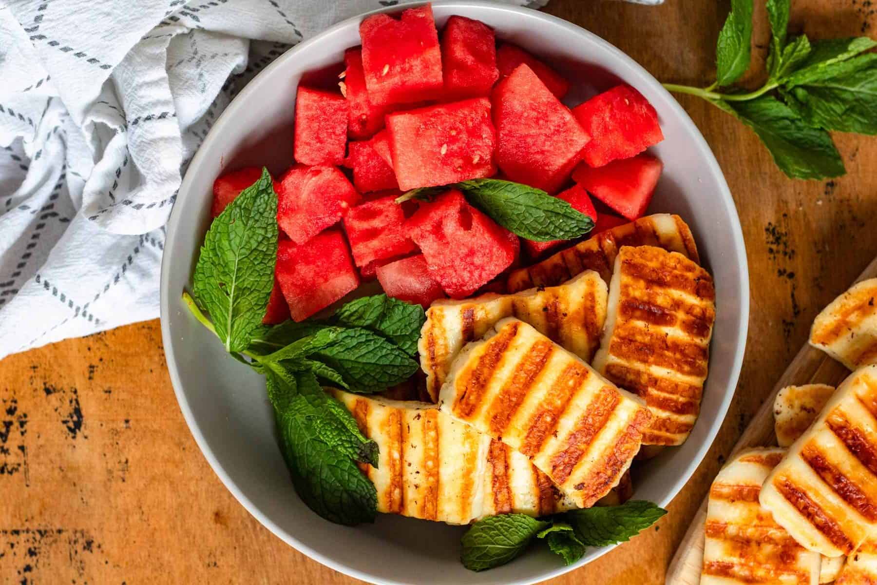 A bowl with grilled halloumi slices, cubed watermelon, and fresh mint leaves on a wooden table—perfect for those seeking fresh recipes to elevate any guest menu.