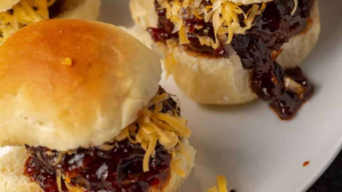 Two barbecue sauce-covered sliders topped with shredded cheese are served on a white plate, making them perfect Game Day Appetizers or Party Snacks.