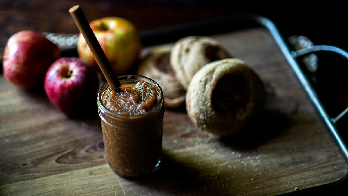 A jar of applesauce with a spoon, two apples, and two English muffins on a wooden tray—perfect for enjoying classic apple dishes during apple season.