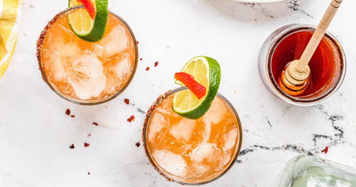 Two iced cocktails, perfect for sipping, are garnished with lime and chili slices and sit on a white surface next to a jar of honey with a honey dipper—bringing a touch of fire to each refreshing glass.
