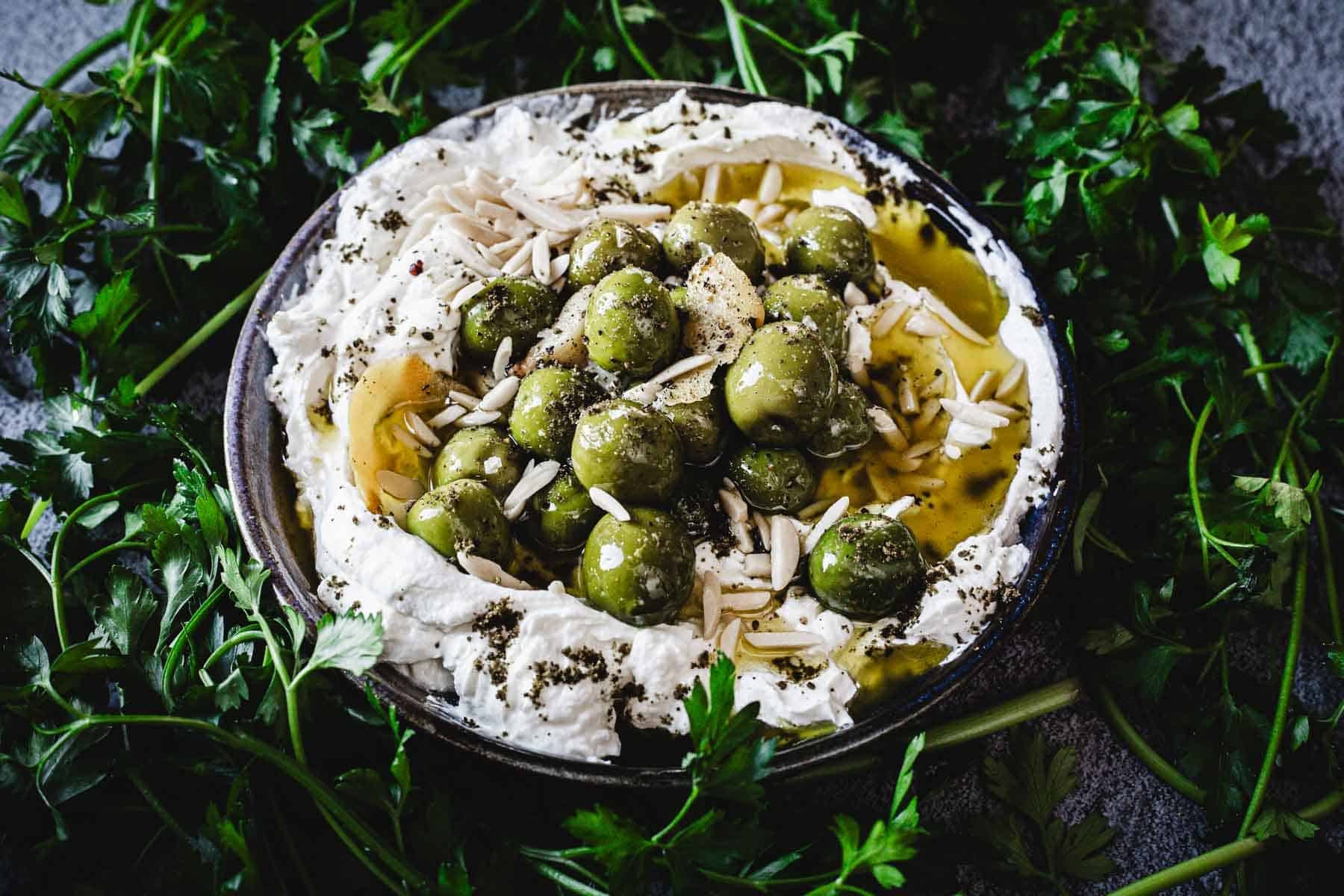 A bowl of labneh topped with green olives, slivered almonds, olive oil, and herbs, surrounded by fresh parsley—perfect for game day snacking or as a delicious addition to your favorite cold dips spread.