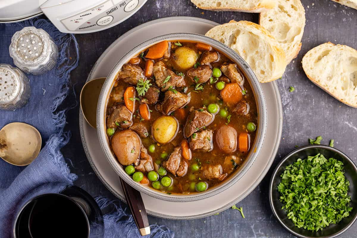 A bowl of beef stew with potatoes, carrots, and peas, served with sliced bread, chopped herbs, a spoon, and a blue napkin on a dark surface—perfect for cozy Fall recipes or as an inviting addition to your collection of Crockpot recipes.