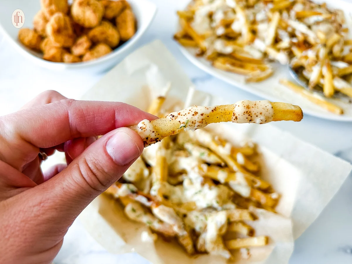 A hand holds a seasoned French fry with white sauce, making it the perfect choice for Game Day Appetizers. Plates of French fries and a bowl of popcorn chicken are in the background on a white surface, ready to be enjoyed as delicious party snacks.