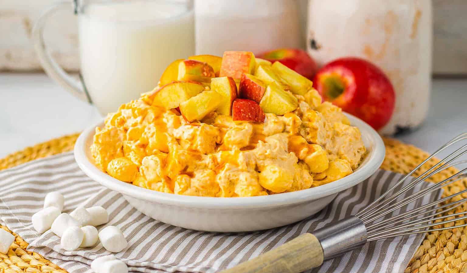 A bowl of orange fruit salad topped with chopped apples, surrounded by marshmallows, a whisk, apples, and a jug of milk on a striped cloth—this inviting scene is perfect inspiration for apple recipes to enjoy during apple season.