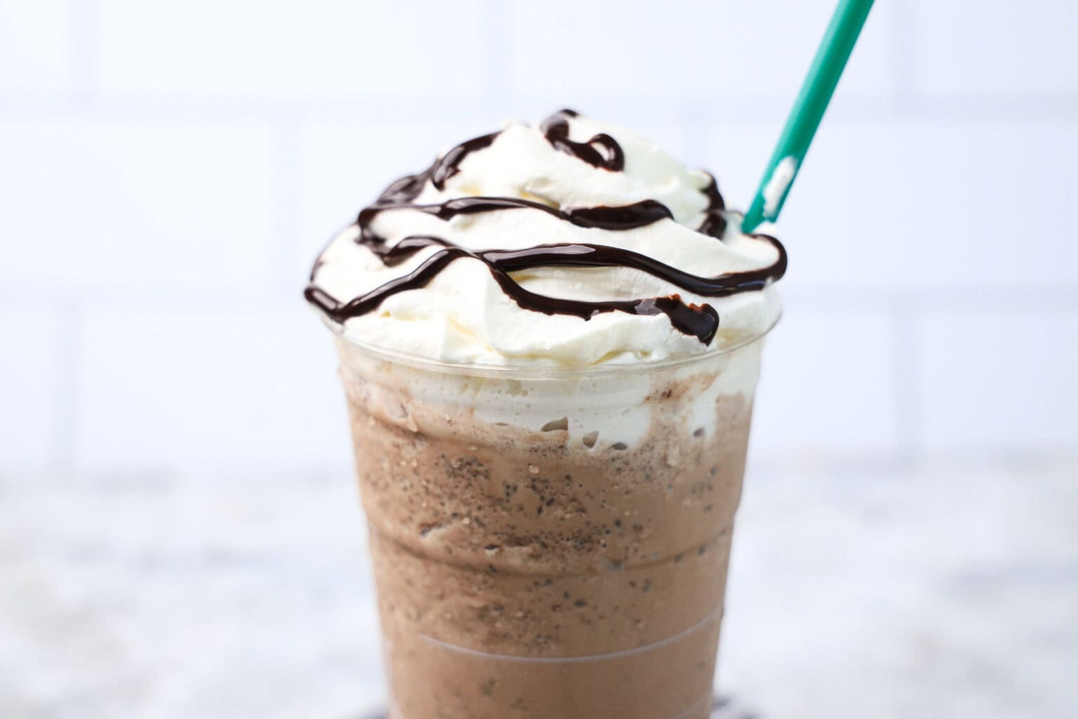 A plastic cup filled with a blended iced coffee drink, topped with whipped cream and chocolate syrup—a true delight for chocolate lovers—served with a green straw.