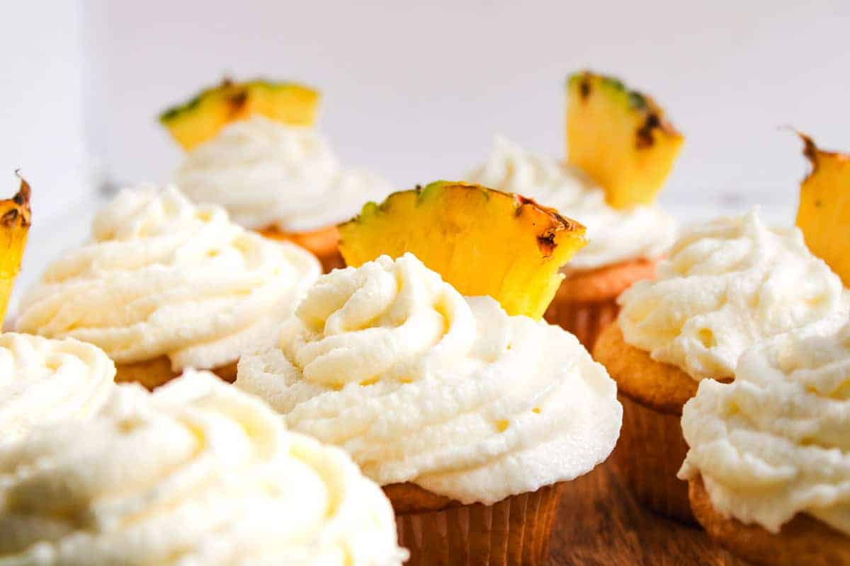 Cupcakes topped with swirls of white frosting and garnished with small slices of pineapple are arranged on a wooden surface, making them perfect easy desserts for any cookout.