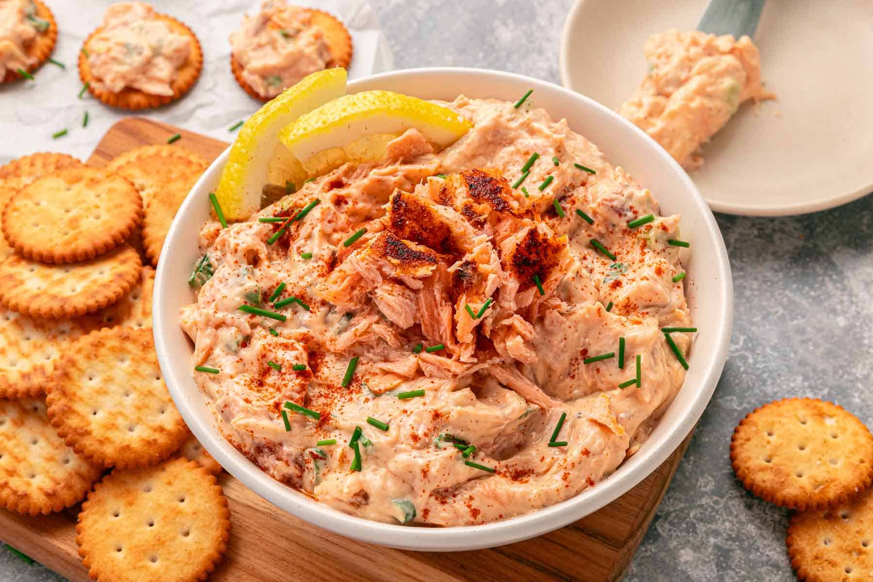A bowl of salmon dip garnished with lemon wedges and chives, surrounded by round crackers on a wooden board—perfect for easy snacking or serving as one of your favorite cold dips at game day gatherings.