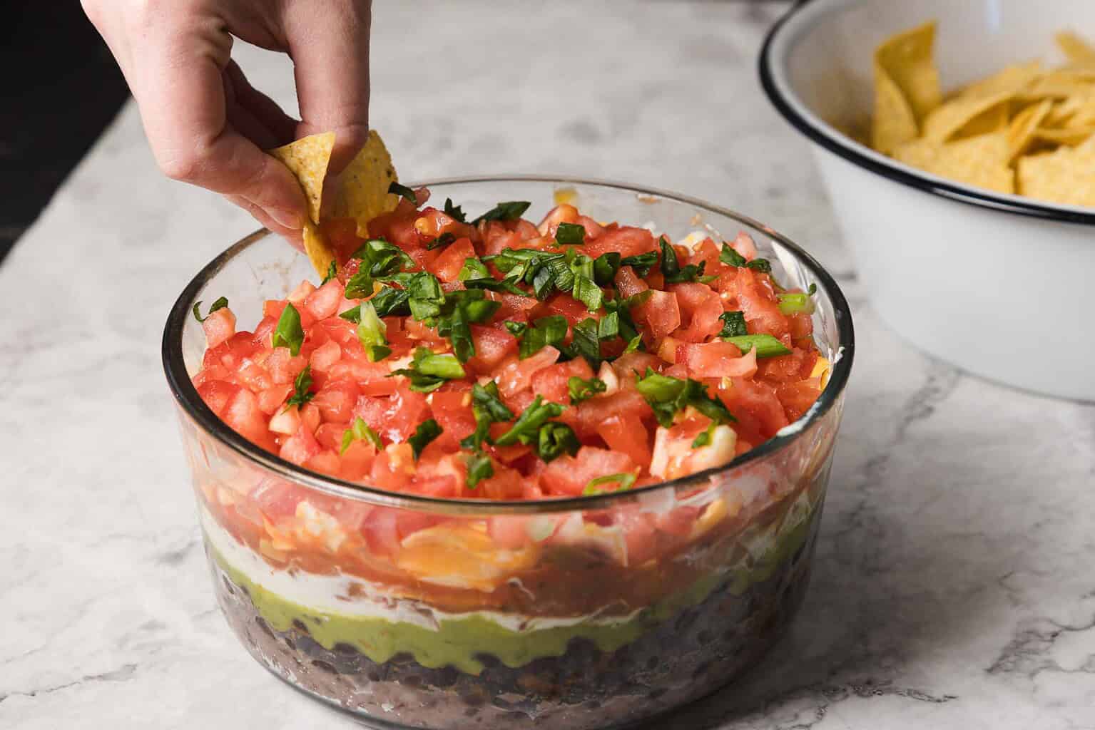 A hand dips a tortilla chip into a glass dish of seven-layer dip with visible layers of beans, guacamole, sour cream, cheese, and tomatoes—perfect for snacking or as one of your favorite game day snacks. A bowl of chips is in the background.
