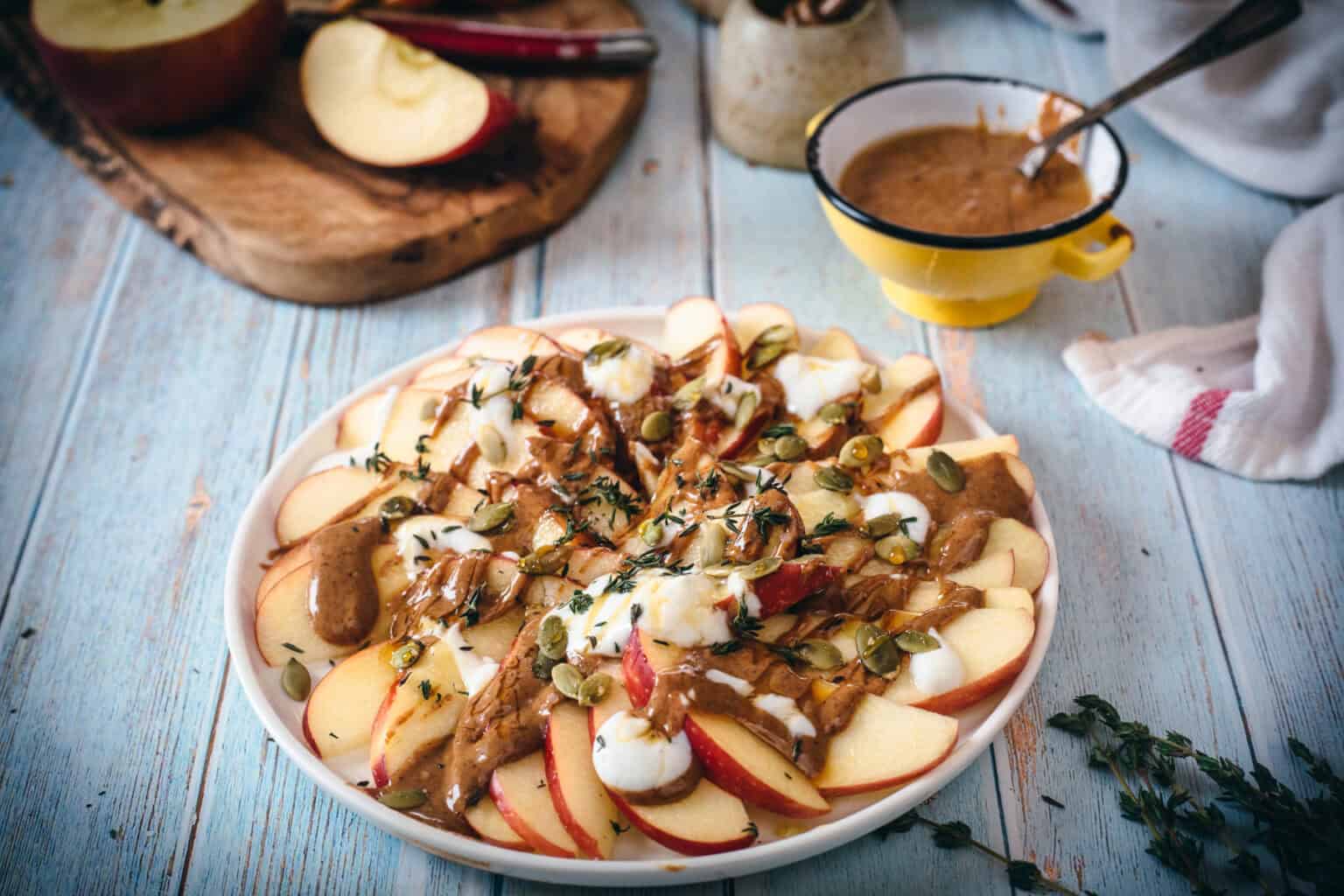 Sliced apples on a plate topped with yogurt, nut butter, seeds, and herbs make a fresh addition to your favorite apple recipes, served alongside a yellow cup of sauce and a cutting board filled with more apple slices in the background. Perfect for celebrating apple season with seasonal recipes everyone will love.
