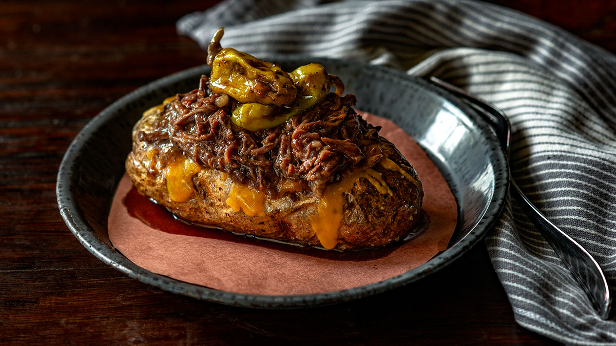 Baked potato topped with shredded beef, melted cheese, and pickled peppers on a black plate with a striped napkin—an easy slow cooker twist on classic comfort food that’s perfect for fall recipes.