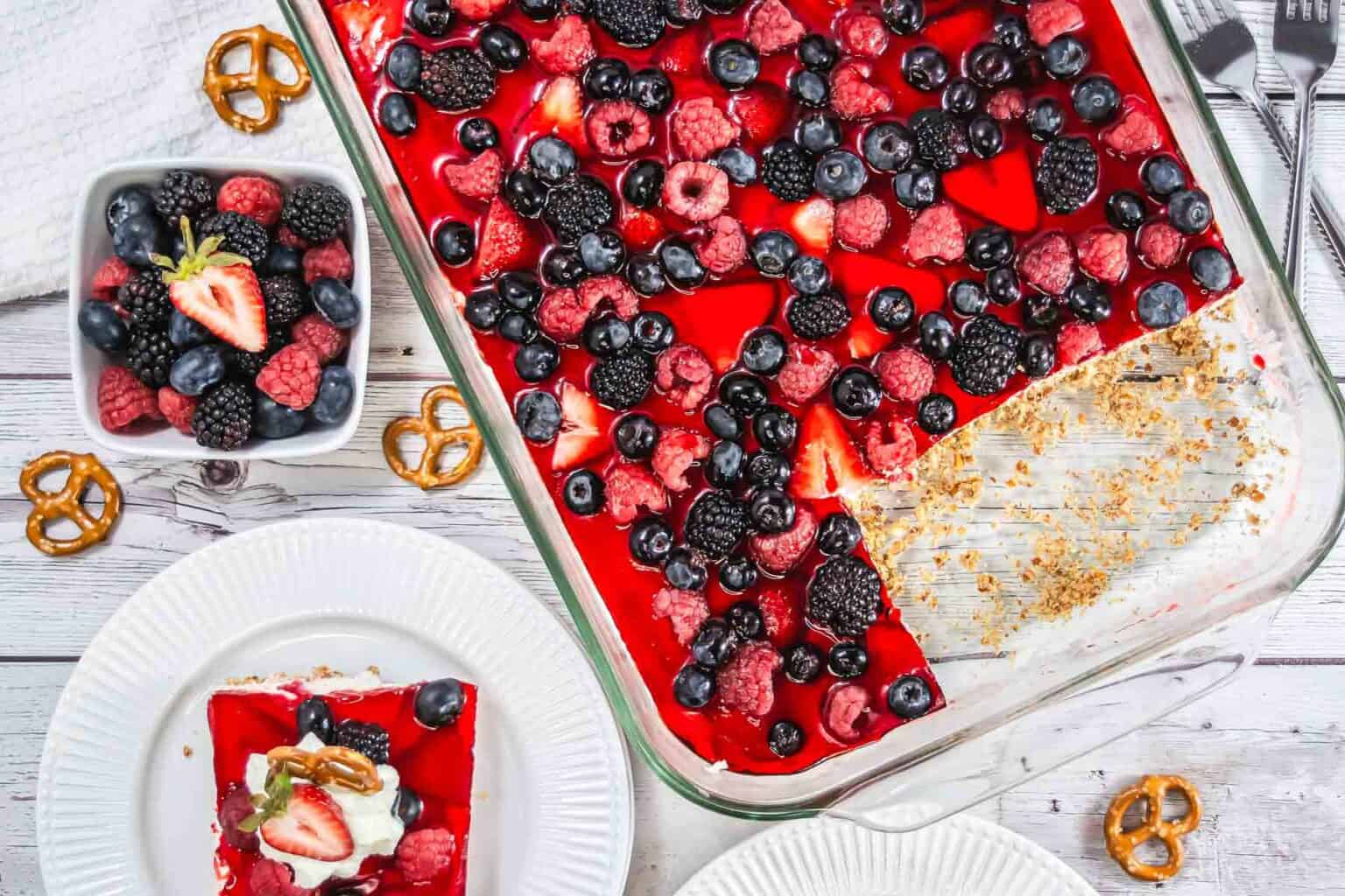A glass dish of berry-topped dessert with a missing corner piece, surrounded by plates with servings, fresh berries, and pretzels on a white wooden table—a delightful option for those searching for summer recipes or cookout ideas to elevate your next Labor Day cookout.