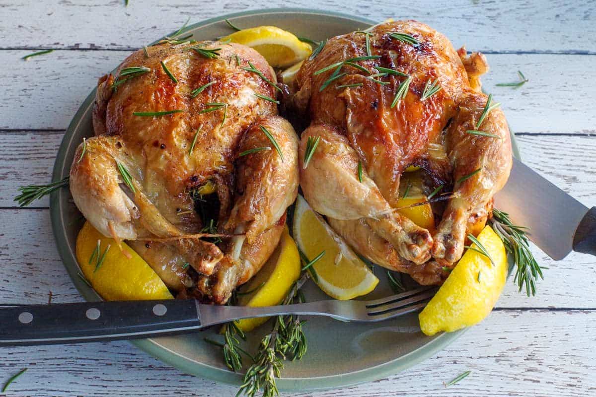 Two roasted Cornish hens garnished with rosemary, served on a plate with lemon wedges and accompanied by a knife and fork—a delightful example of new chicken dishes for those seeking innovative roasted chicken recipes.