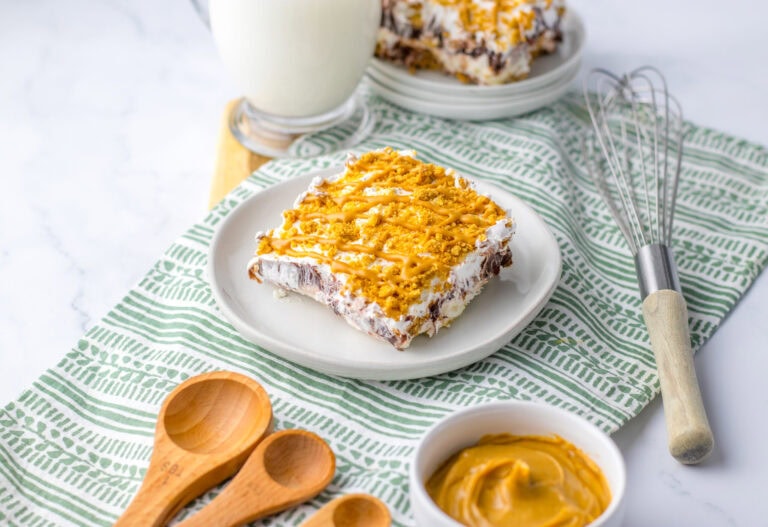 A dessert bar with a creamy topping and orange drizzle sits on a white plate, surrounded by a glass of milk, whisk, wooden spoons, and a bowl of spread on a patterned cloth—a delicious addition to your collection of peanut butter desserts.