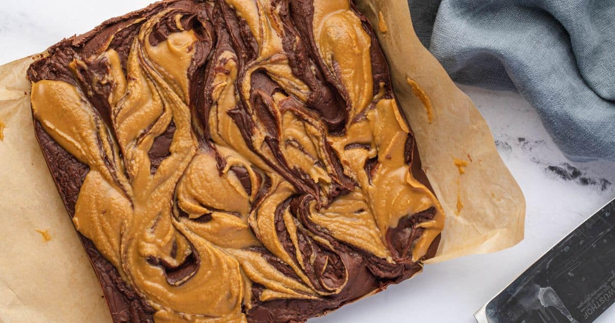 A tray of brownies with a swirled peanut butter topping on parchment paper, placed next to a gray cloth and a metal spatula—perfect for anyone searching for irresistible peanut butter desserts or new dessert recipes to try.