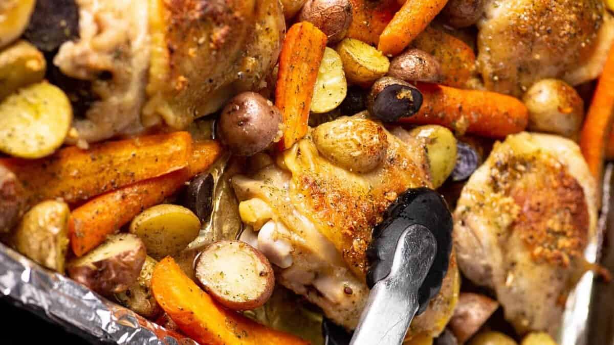 Sheet pan with roasted chicken thighs, baby potatoes, and carrots, seasoned and cooked to perfection—an ideal choice for easy dinners. Metal tongs are picking up a piece from this delicious sheet pan meal, making it perfect for satisfying weeknight meals.