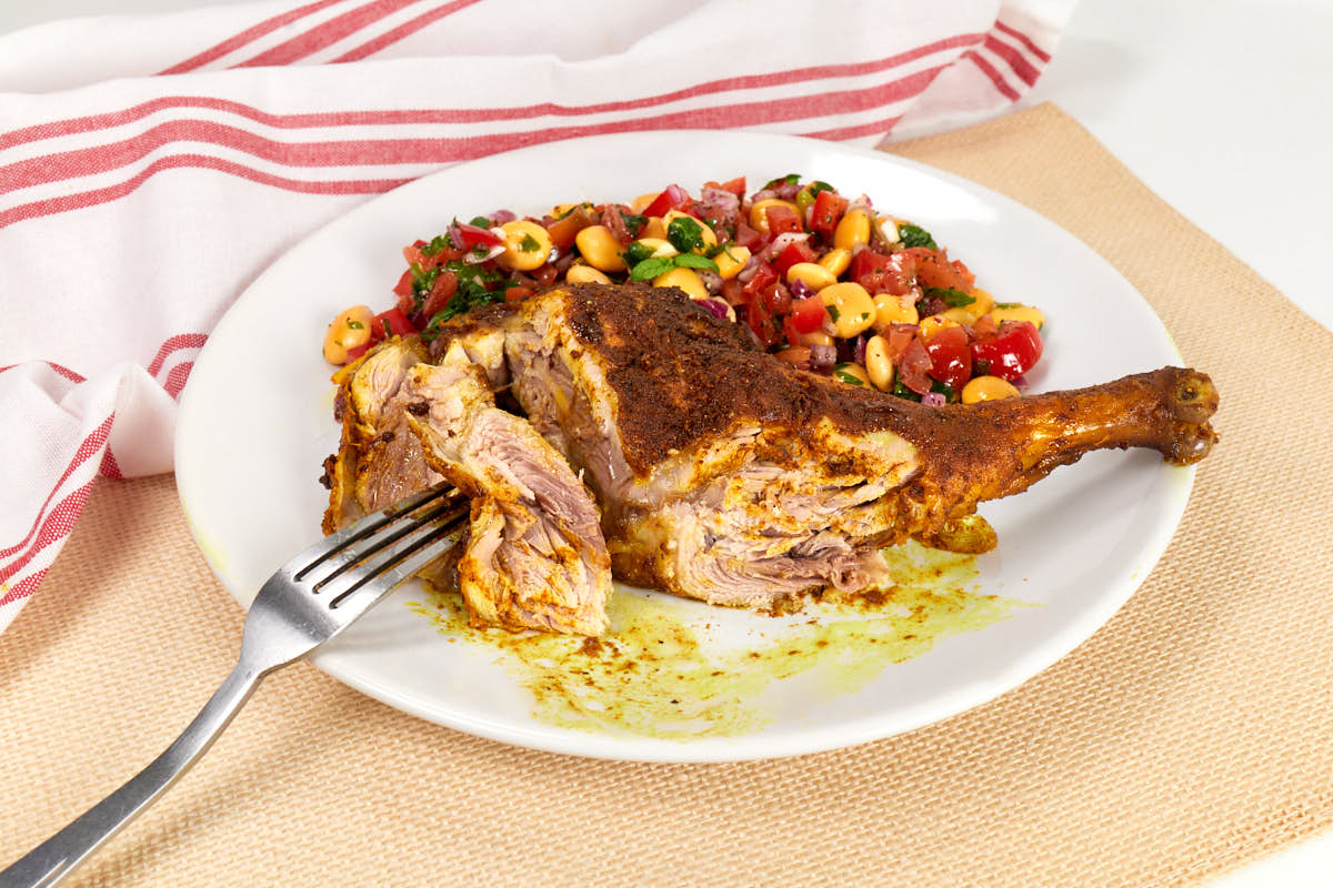 A seasoned roasted chicken leg on a white plate with a bean and tomato salad, set on a beige placemat with a red-striped cloth in the background—a delicious showcase for those seeking roasted chicken recipes.