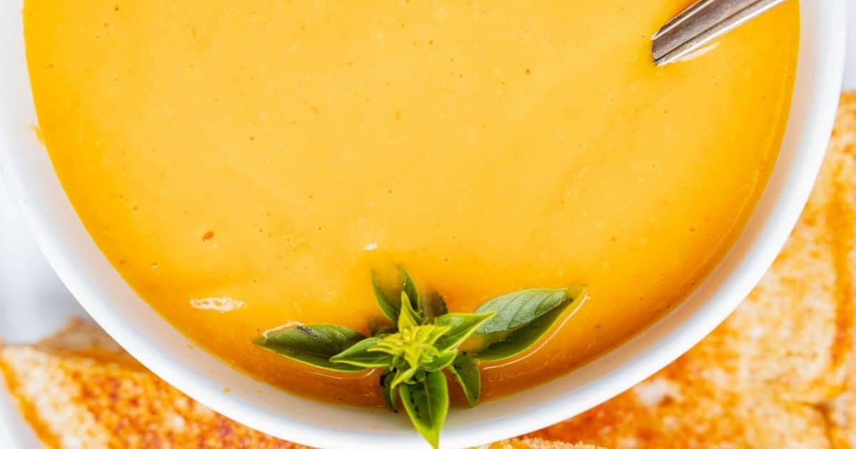 A bowl of creamy orange soup—perfect for cozy autumn meals—garnished with fresh basil leaves, with a spoon and toasted bread on the side.