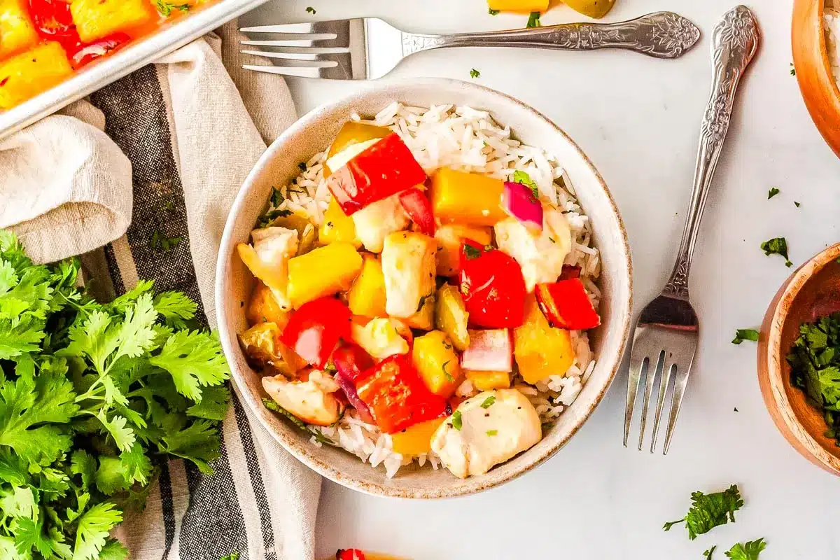 Bowl of white rice topped with colorful chunks of chicken, red and yellow bell peppers, and onions, with herbs scattered nearby—an inviting option for easy dinners. Forks and a napkin are placed beside the bowl.