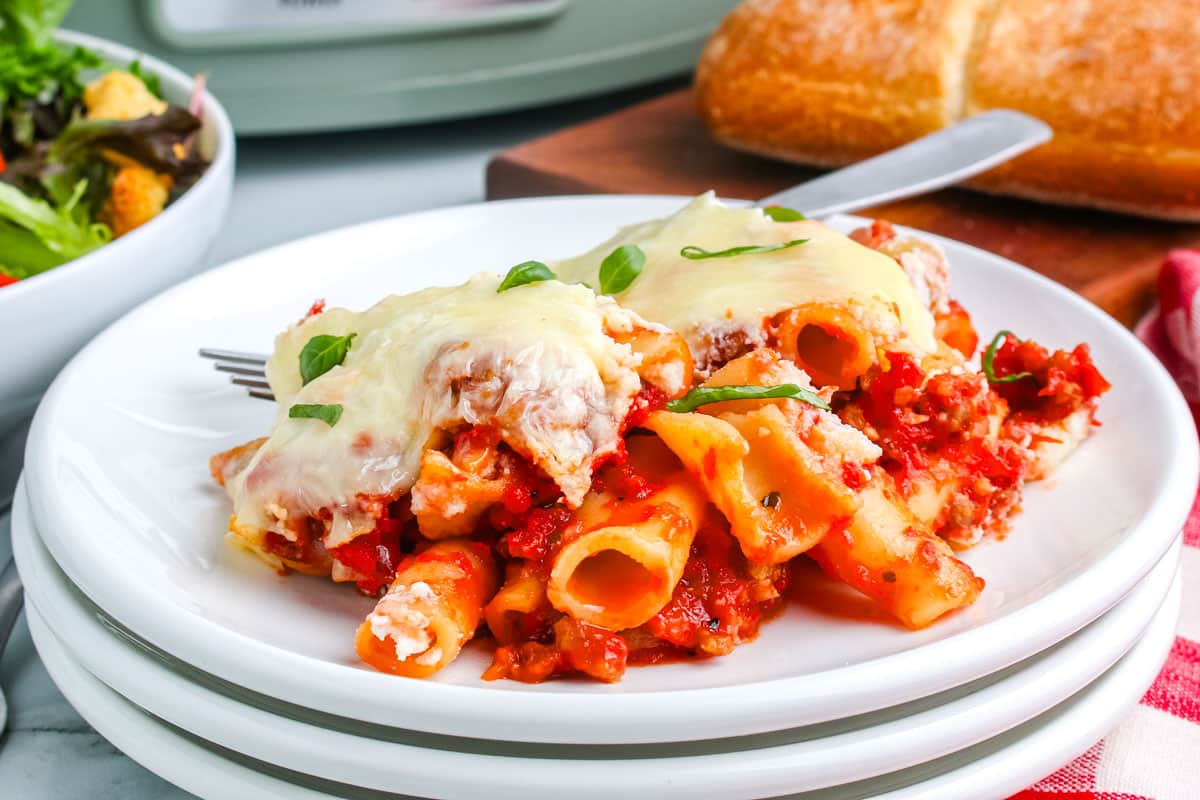 Baked ziti pasta with tomato sauce, melted cheese, and herbs served on a white plate with a fork; bread and salad are in the background—perfect for cozy fall recipes or easy slow cooker meals.