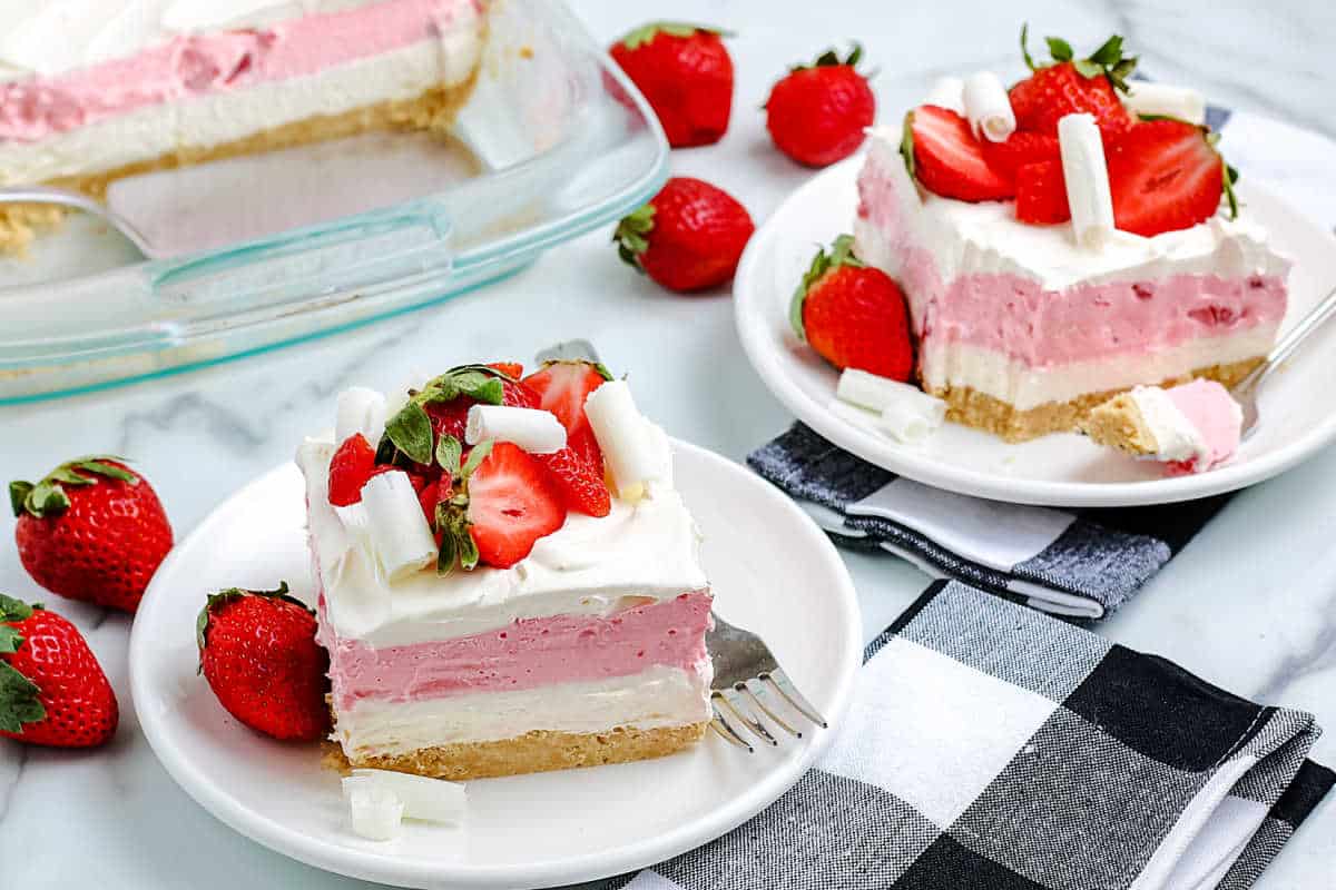 Two plates with layered strawberry dessert bars topped with whipped cream, fresh strawberries, and white chocolate curls, next to a baking dish and whole strawberries on a marble surface—these easy desserts are perfect for cookouts or festive gatherings like Labor Day.