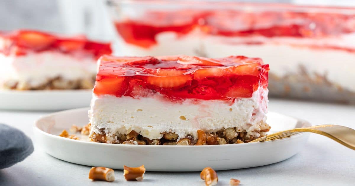 A slice of strawberry pretzel dessert with a pretzel crust, creamy middle layer, and strawberry gelatin topping, served on a white plate with a fork. Other pieces are visible in the background—making it perfect for potlucks and beloved among tried-and-true recipes for any gathering.