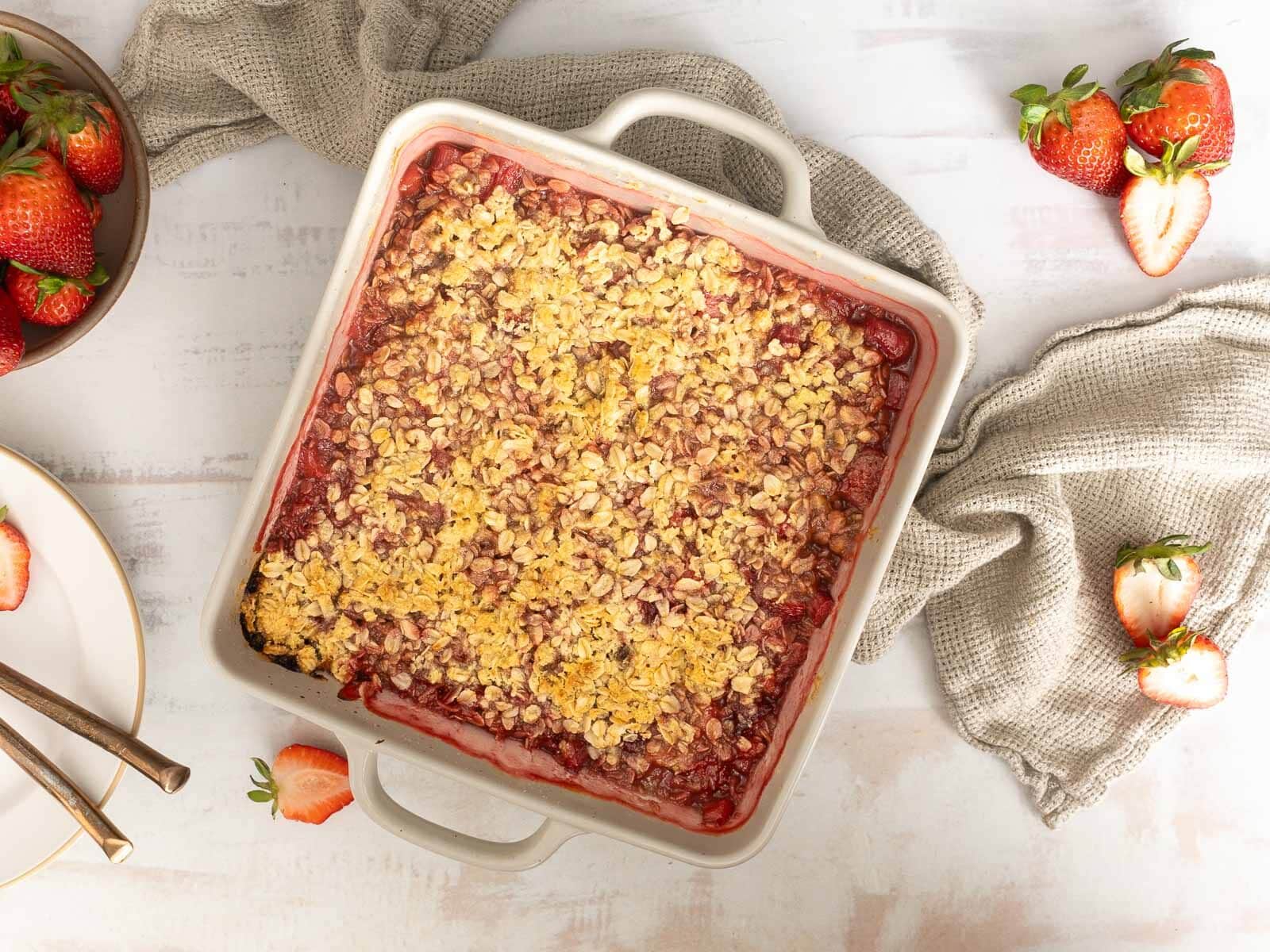 A square baking dish filled with strawberry crumble sits on a light surface, surrounded by fresh strawberries, napkins, and a plate with forks—an irresistible centerpiece for summer desserts or anyone seeking delicious gluten free treats.