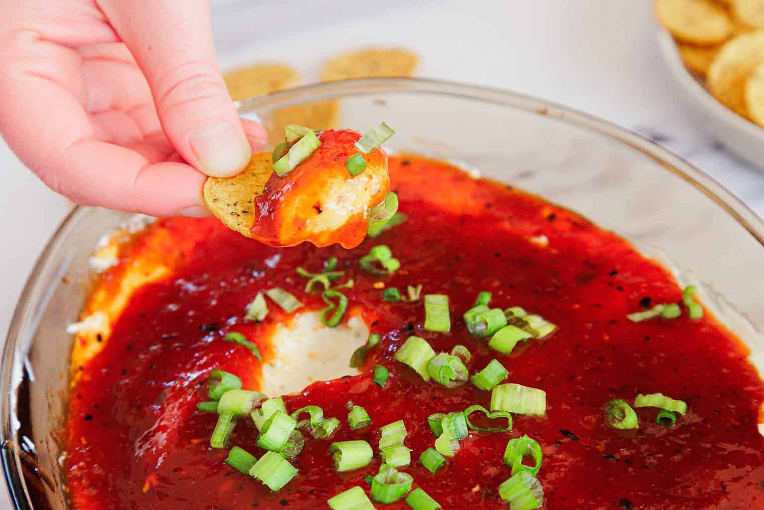 A hand dips a round chip into a glass dish filled with a layered dip topped with red sauce and chopped green onions—perfect for game day snacks or party appetizers.