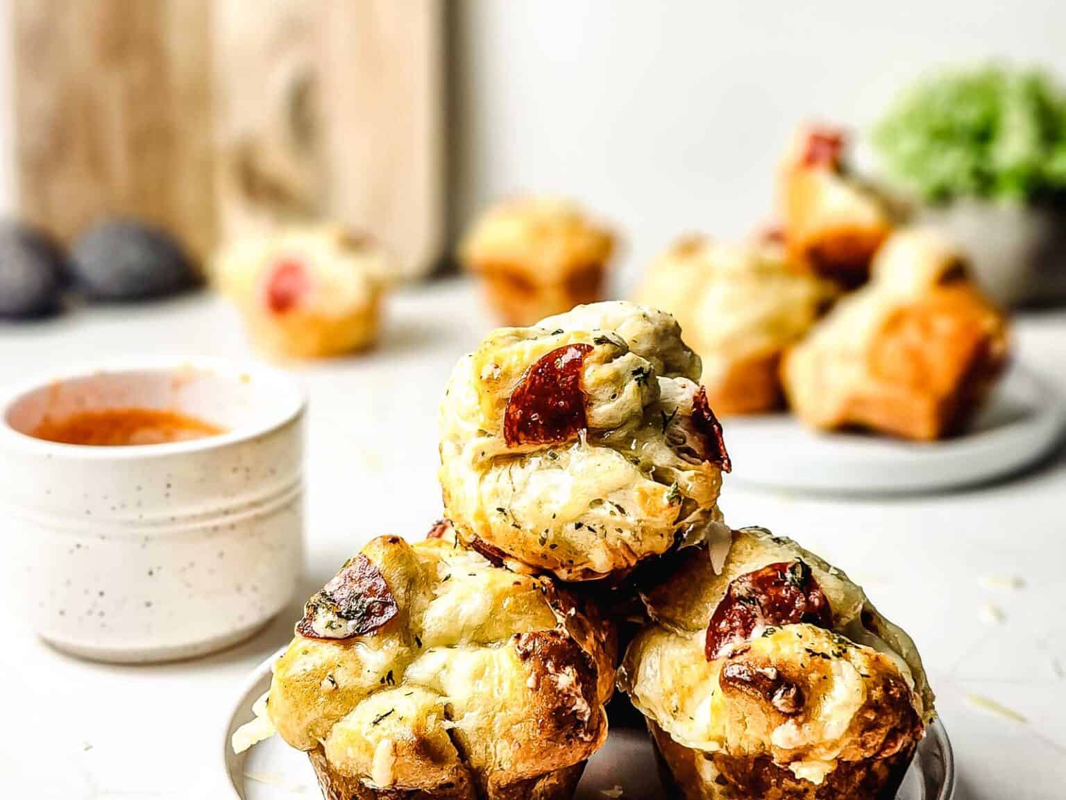 Three savory muffins with melted cheese and pepperoni are stacked on a plate, making the perfect Game Day Appetizers, with a cup of dipping sauce nearby and more muffins and greens in the background.