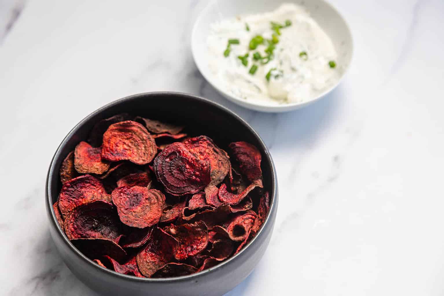 A bowl of beet chips sits on a white surface next to a small bowl of creamy dip garnished with chopped green herbs, making it a vibrant addition to your favorite vegetable dishes or vegetarian recipes.
