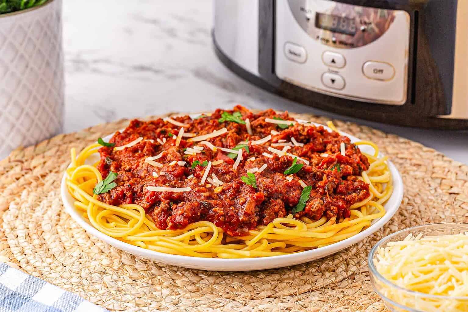 Plate of spaghetti topped with meat sauce and shredded cheese, placed on a woven mat, with a slow cooker and a bowl of cheese in the background—a cozy scene perfect for fall recipes and inspired by classic Crockpot recipes.