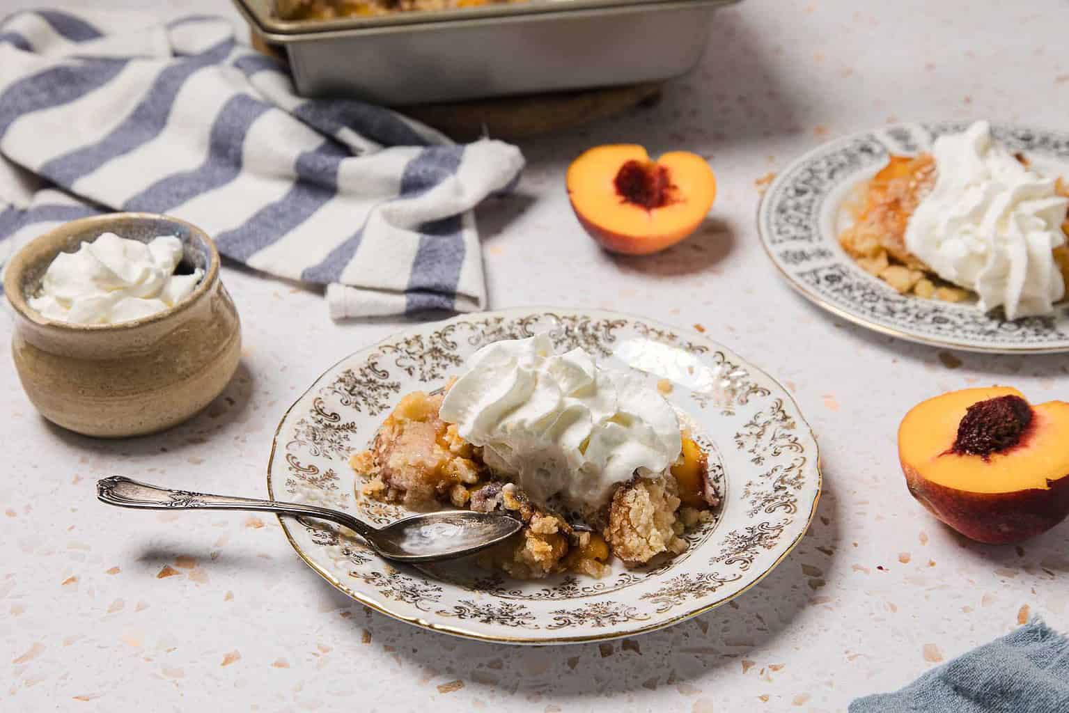 A plate and bowl of peach cobbler topped with whipped cream, with fresh peach halves and a striped towel in the background—perfect for easy desserts or Labor Day desserts at your next cookout.