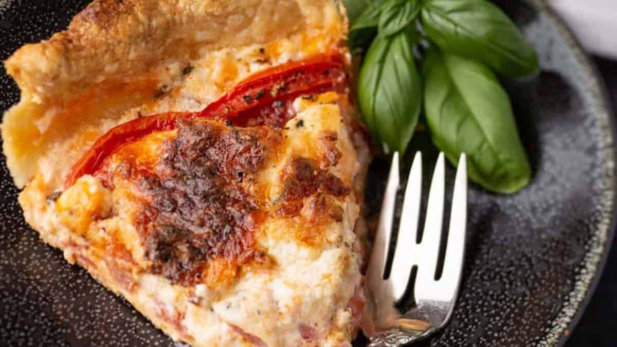 A slice of tomato and cheese quiche on a dark plate with a fork and fresh basil garnish—perfect for those seeking delicious summer recipes or looking to highlight their favorite garden tomato recipes.