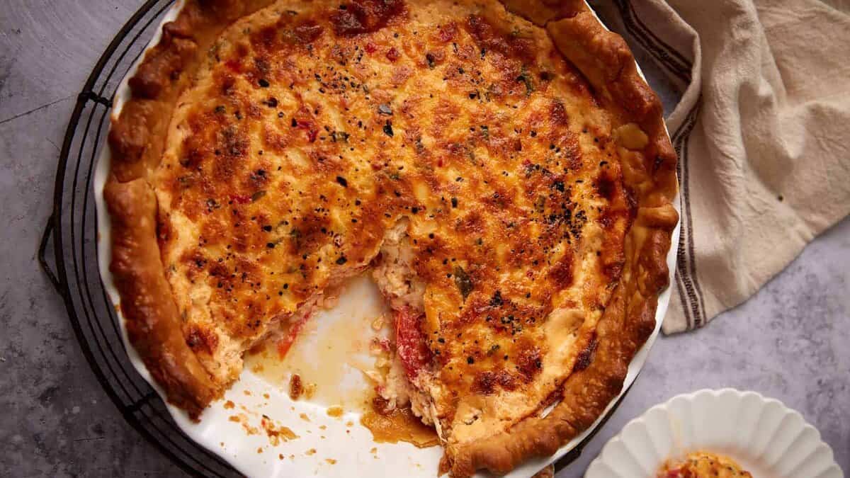 A baked tomato pie with a golden-brown crust in a white dish, one slice removed and placed on a nearby plate—this make-ahead recipe is perfect for stress-free hosting as a delicious addition to your Labor Day sides.