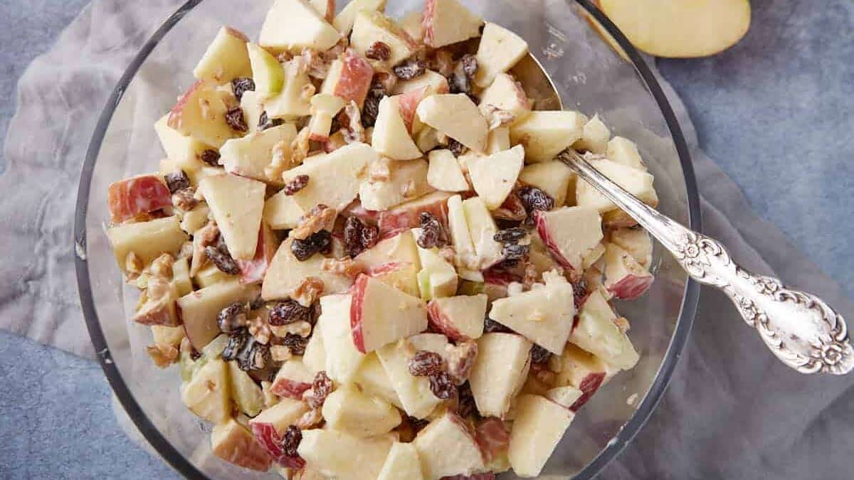 Glass bowl filled with chopped apples, raisins, and walnuts, mixed with a light creamy dressing—perfect for those craving refreshing apple dishes during apple season. A silver serving spoon rests in the bowl.