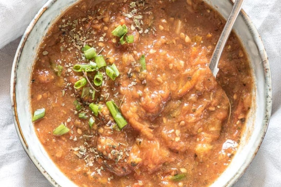 A bowl of thick, brownish tomato-based soup or stew garnished with chopped green onions and herbs, with a spoon inside.