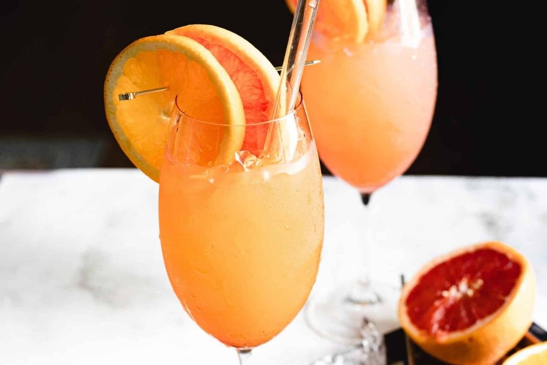 Two glasses of orange-pink cocktail garnished with grapefruit and orange slices, each with a silver straw, sit on a white surface—perfect summer refreshments for those seeking cool drinks to enjoy on Labor Day.
