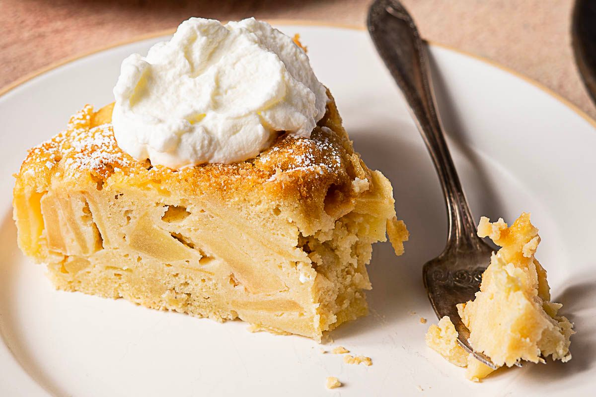A slice of apple cake with whipped cream on top sits on a white plate next to a fork and a small piece of cake, making it the perfect treat to enjoy during apple season or when exploring new apple recipes.