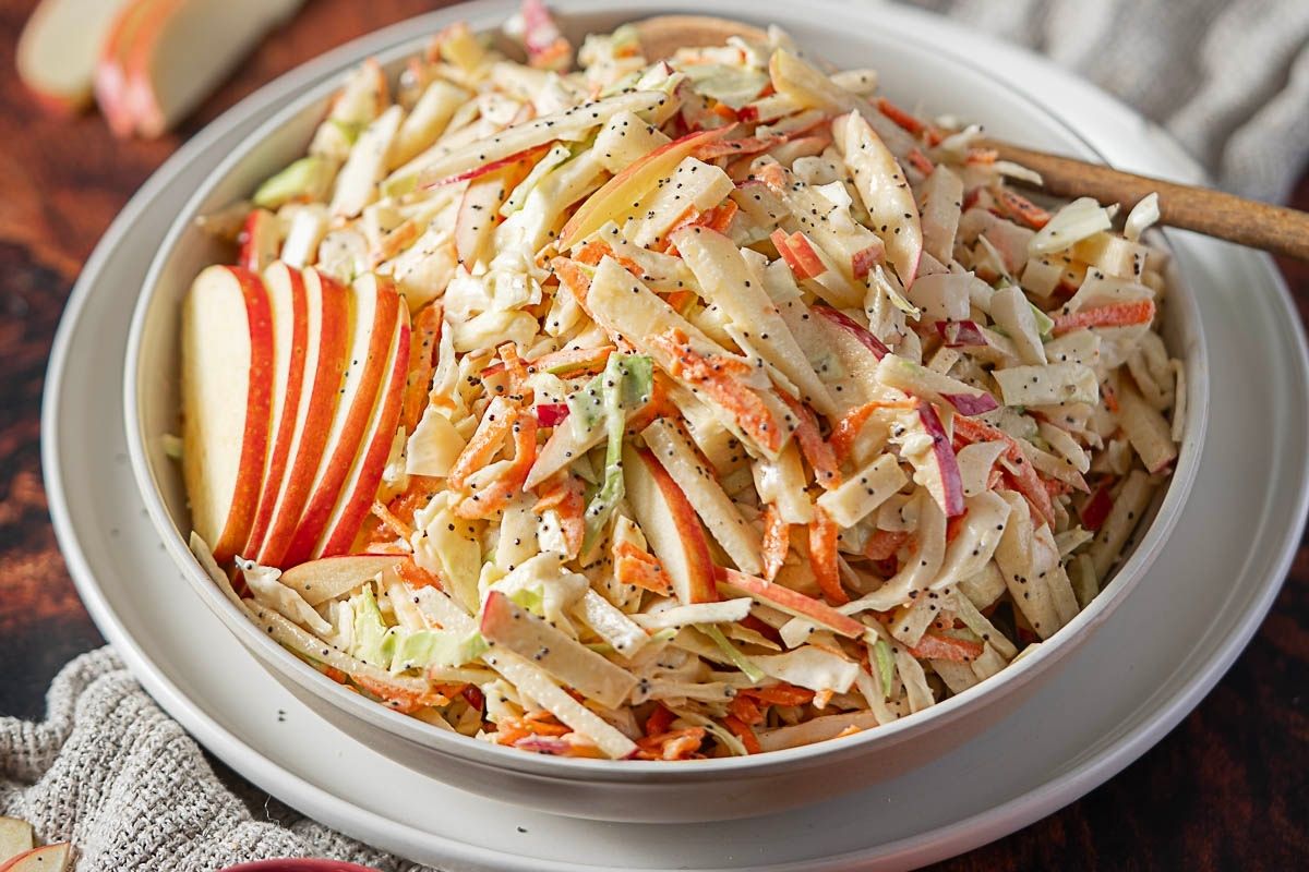A bowl of coleslaw with shredded cabbage, carrots, and apple slices, topped with a creamy dressing and poppy seeds, sits on a plate with a wooden spoon—one of those tried-and-true recipes that always shines among popular potluck dishes.