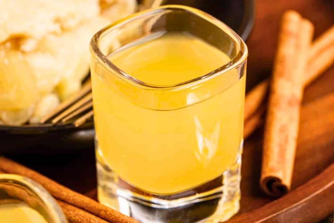 A clear glass filled with yellow liquid is on a wooden surface next to two cinnamon sticks, evoking the warmth of apple season, with food and a black plate partially visible in the background.