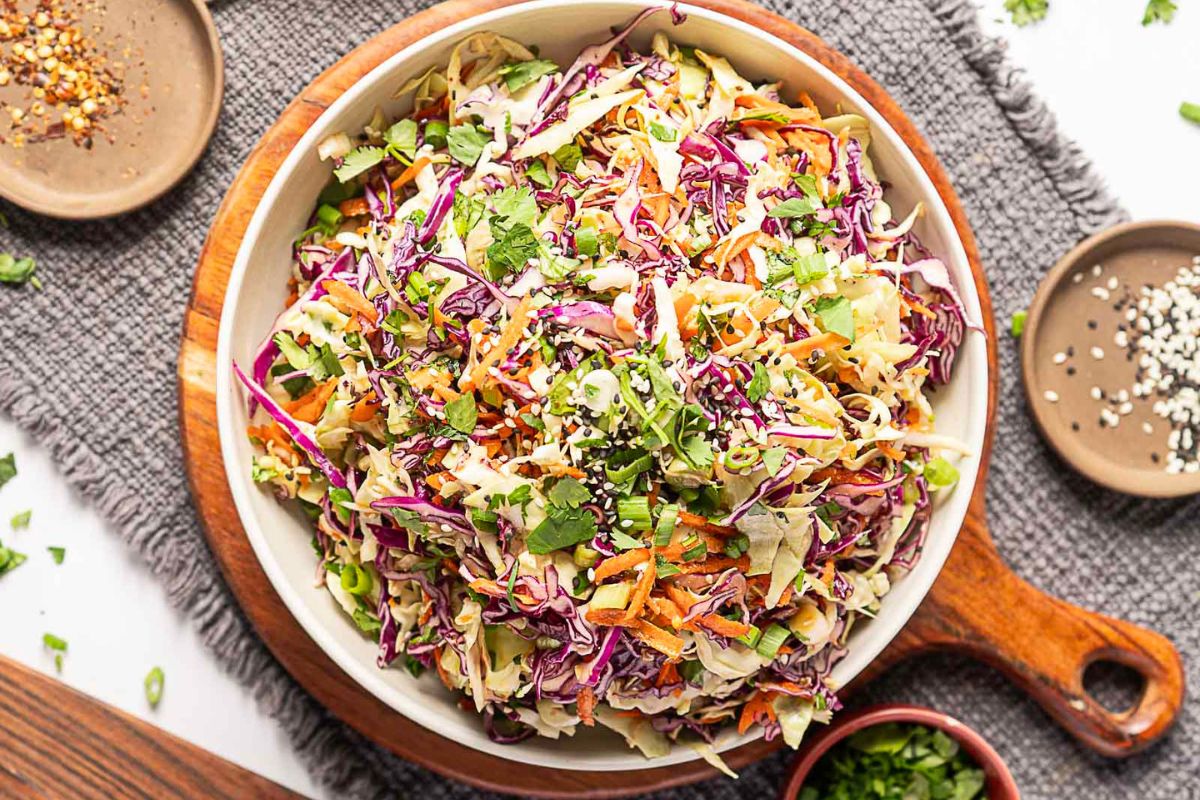A bowl of coleslaw with shredded green and purple cabbage, carrots, chopped herbs, and sprinkled sesame seeds, placed on a gray cloth with small condiment dishes nearby—this make-ahead dish is perfect for stress-free hosting or serving as one of your favorite Labor Day sides.