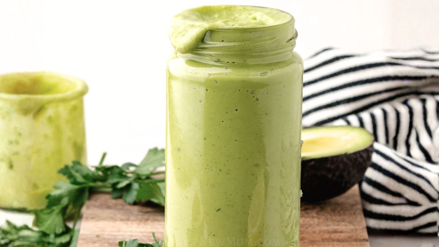 A glass jar filled with green creamy sauce or homemade dressing, with some dripped over the rim. A halved avocado, herbs, and a striped cloth are in the background—perfect inspiration for your next salad dressing recipe.
