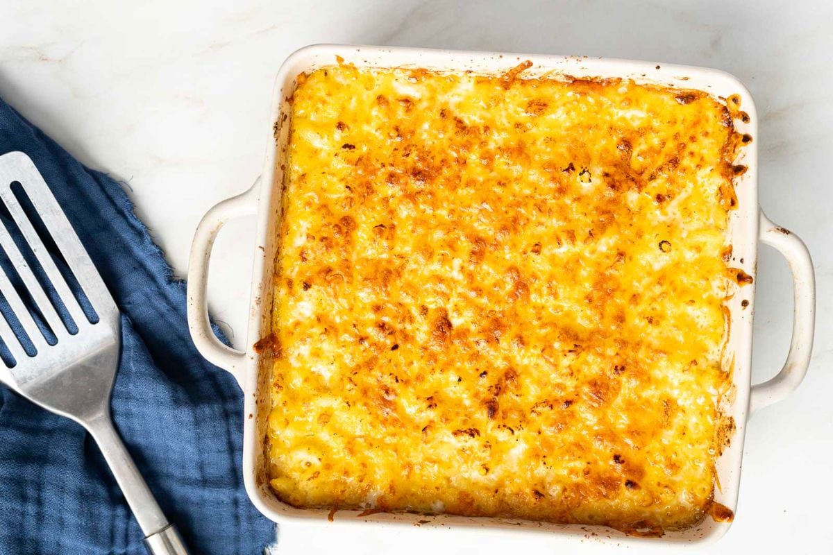 A square baking dish filled with baked macaroni and cheese with a golden, crispy top sits next to a metal spatula and a blue cloth—an ideal make-ahead option for stress-free hosting, perfect for serving as one of your Labor Day sides.