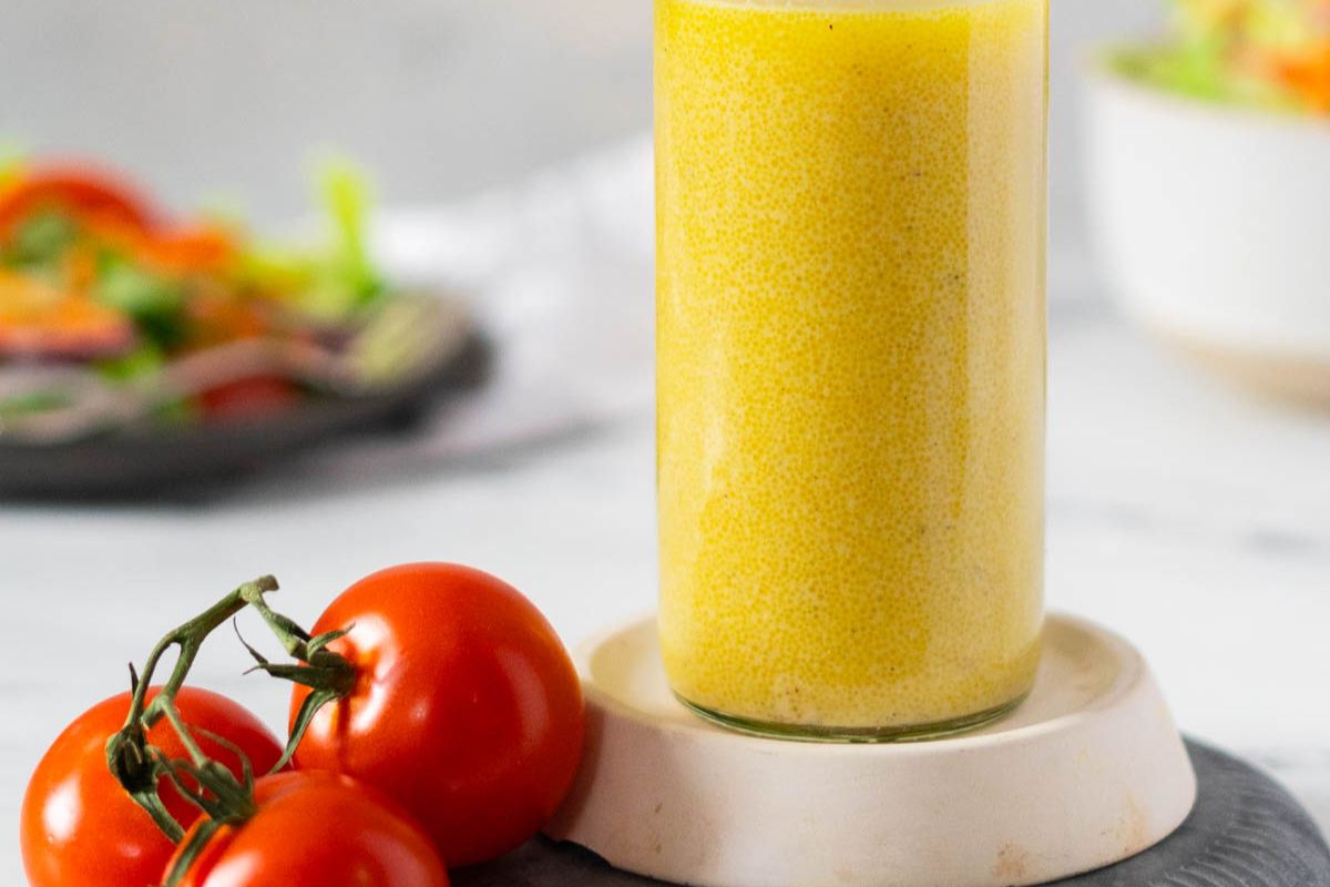 A glass of yellow salad dressing sits on a small plate next to three vine tomatoes, with a blurred salad in the background—perfect inspiration for anyone seeking homemade dressings to elevate their meals.
