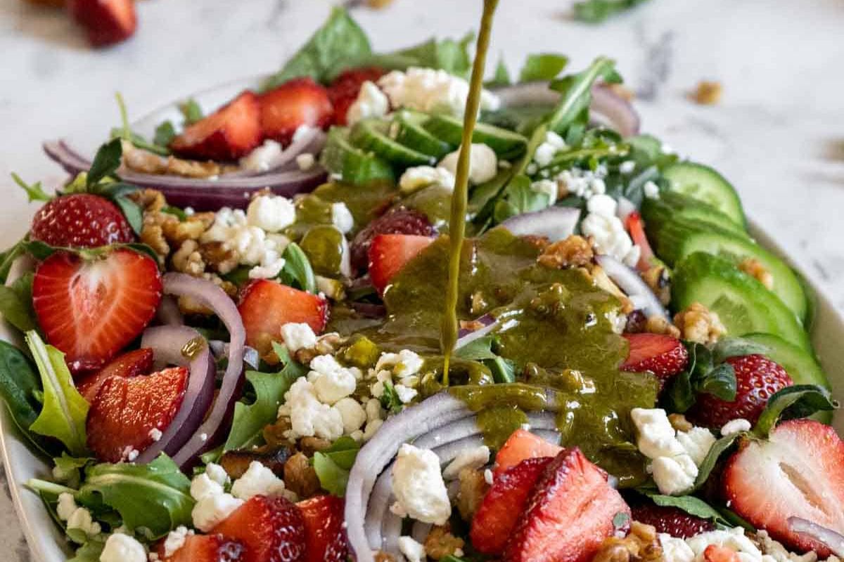 A salad with sliced strawberries, cucumber, red onions, walnuts, and feta cheese is being drizzled with one of the best homemade dressings—a perfect addition for those who love unique salad dressings and homemade recipes.