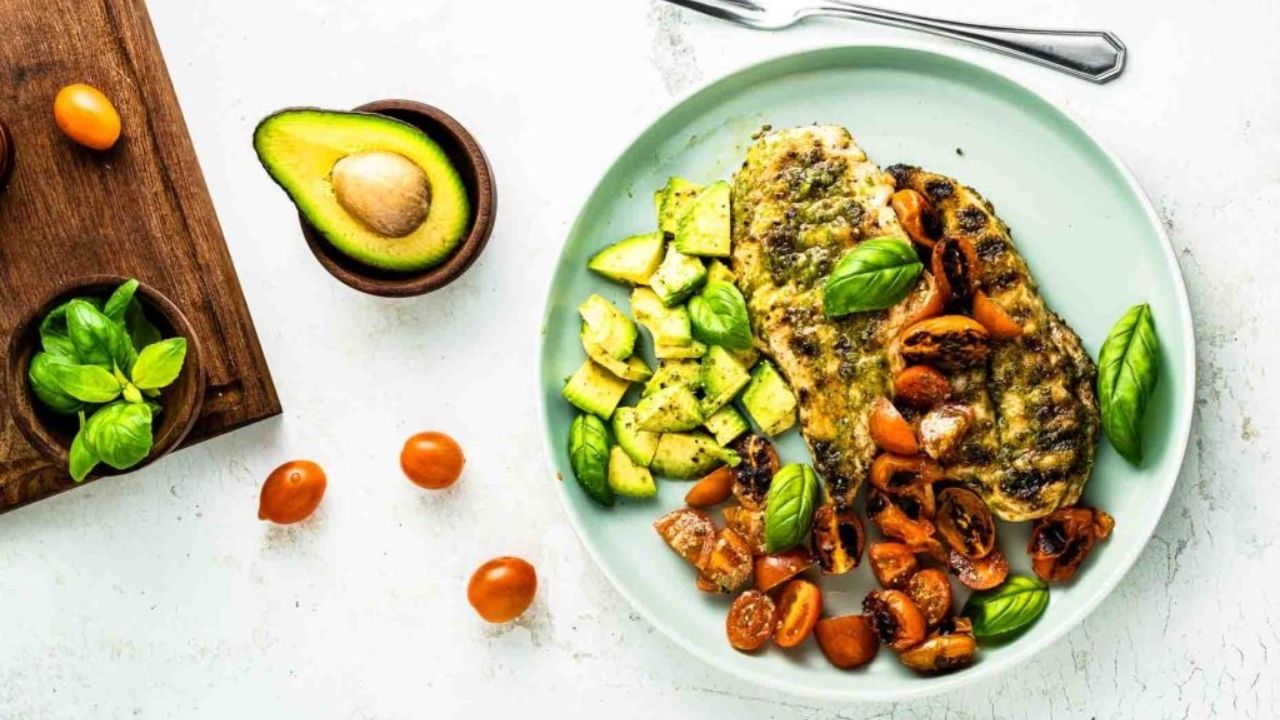 A plate with grilled fish, diced avocado, and sliced cherry tomatoes garnished with basil showcases the vibrant flavors of fresh garden meals. A fork, half an avocado, tomatoes, and fresh basil are nearby on the table.